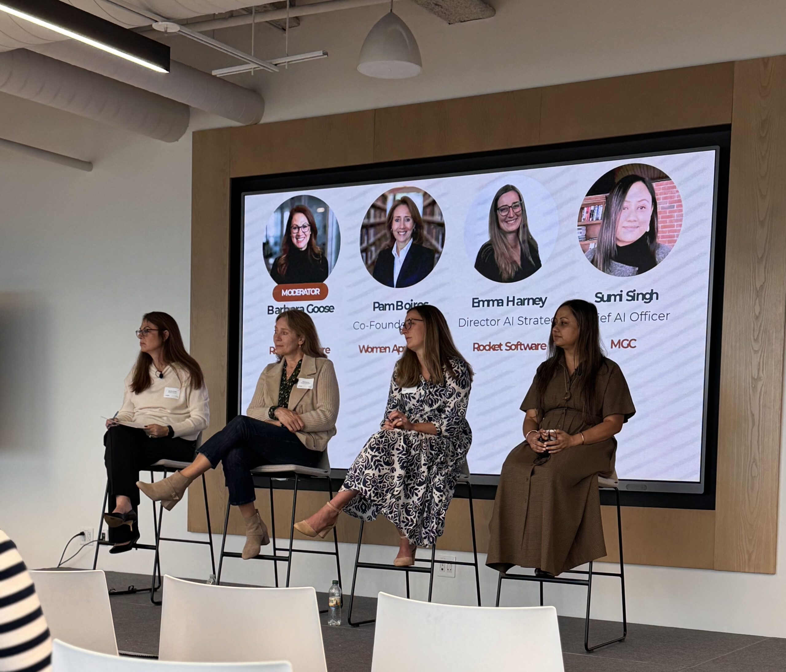 Four panelists seated on stools in front of a screen showing speaker photos at the Boston AI Week panel at Rocket Software